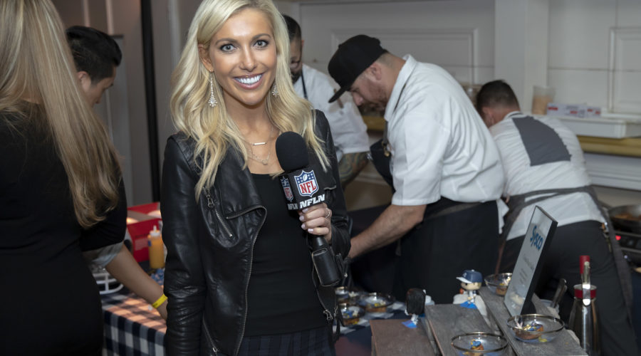 A smiling woman holding an NFL microphone stands in front of a table where chefs prepare food at a culinary event benefiting the Los Angeles Regional Food Bank. She wears a black jacket, exuding enthusiasm as she captures the vibrant atmosphere.