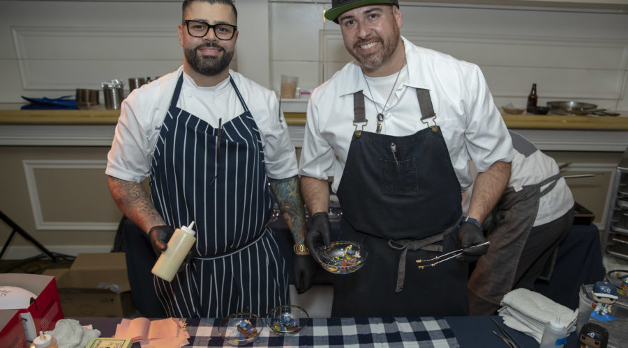 Two chefs wearing aprons stand at a table brimming with cooking ingredients, supporting the Los Angeles Regional Food Bank. One chef holds a squeeze bottle, the other tweezers. They smile at the camera, surrounded by dishes and utensils on a blue checkered tablecloth.