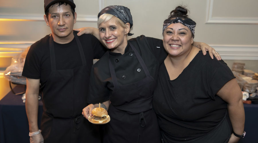 Three people in black attire smile at the camera. The person in the middle holds a small burger, showcasing their culinary skills. Dressed in chefs' uniforms, including aprons and bandanas or caps, they stand proudly before a table of food items at the Los Angeles Regional Food Bank.