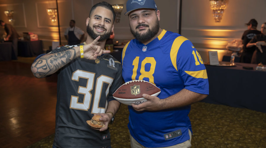 Two men in NFL jerseys stand indoors, one holding a football, as they pose for the camera with friendly gestures. The room is dimly lit with warm lighting and ornate wall fixtures, creating a cozy atmosphere reminiscent of a Los Angeles Regional Food Bank fundraiser.