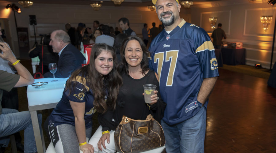 A family of three is smiling at a social event supporting the Los Angeles Regional Food Bank. The man and young girl wear matching sports jerseys. The woman, in black, holds a drink and carries a brown handbag. They're standing by a high table in a room with chandelier lighting.