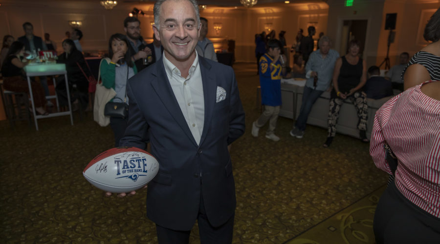 A man in a suit holds a football inscribed with "Taste of the NFL," supporting the Los Angeles Regional Food Bank. In the background, people gather at a social event in a well-lit room adorned with chandeliers.