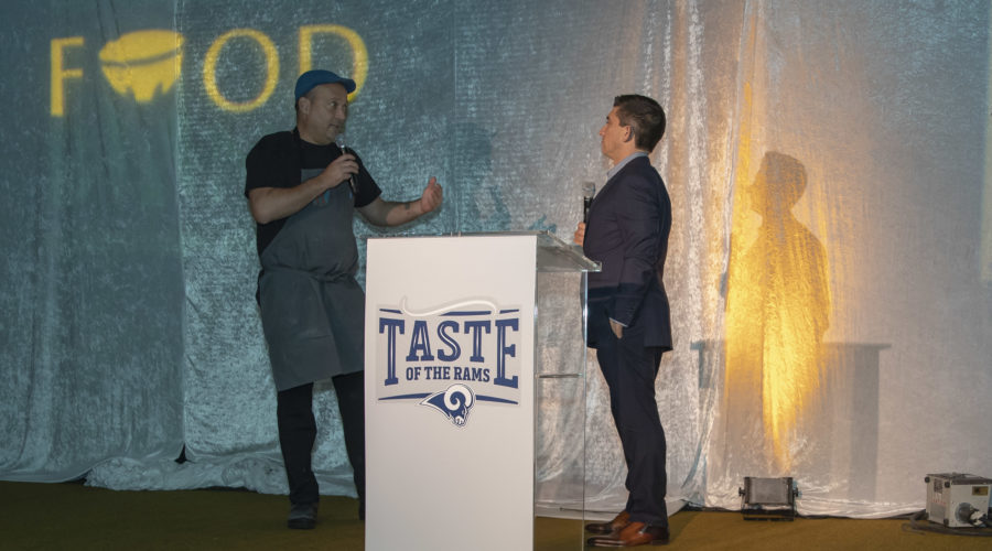 A man in a black shirt and apron speaks into a microphone behind a podium featuring the "Taste of the Rams" logo, proudly supporting the Los Angeles Regional Food Bank. Another man in a suit stands to the side, listening intently. A "FOOD" sign is partially visible on the wall behind them.