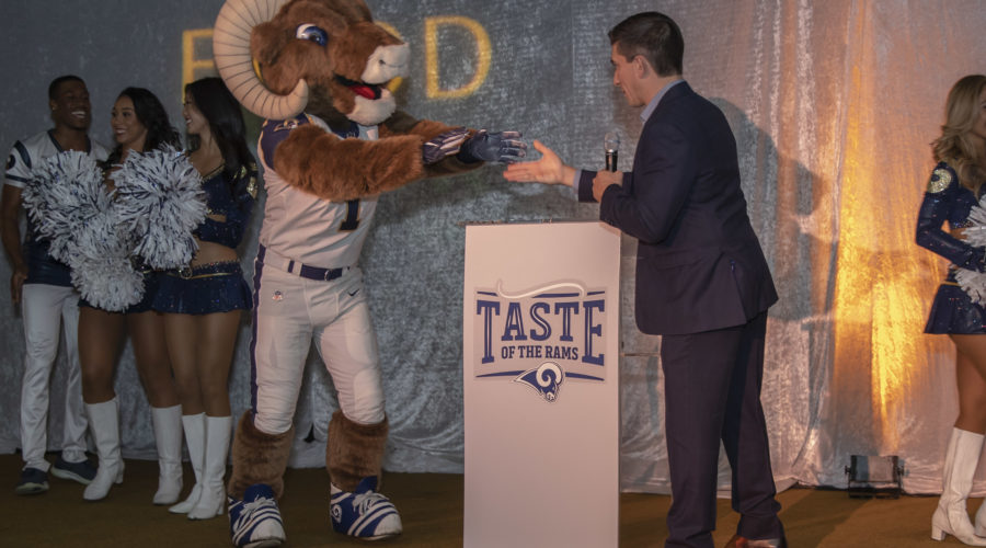 A man in a suit hands a microphone to a costumed ram mascot at a podium emblazoned with "Taste of the Rams," supporting the Los Angeles Regional Food Bank. Cheerleaders stand in the background against a shimmering silver curtain, adding to the festive atmosphere.