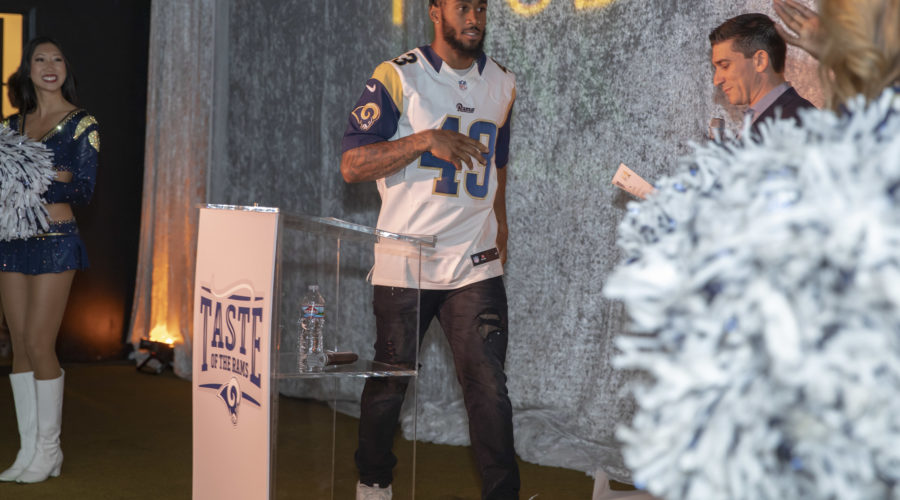 A football player in a Rams jersey stands on stage near a podium at an event for the Los Angeles Regional Food Bank. A cheerleader and another person are visible, clapping, with a silver curtain backdrop displaying the word "FOOD.