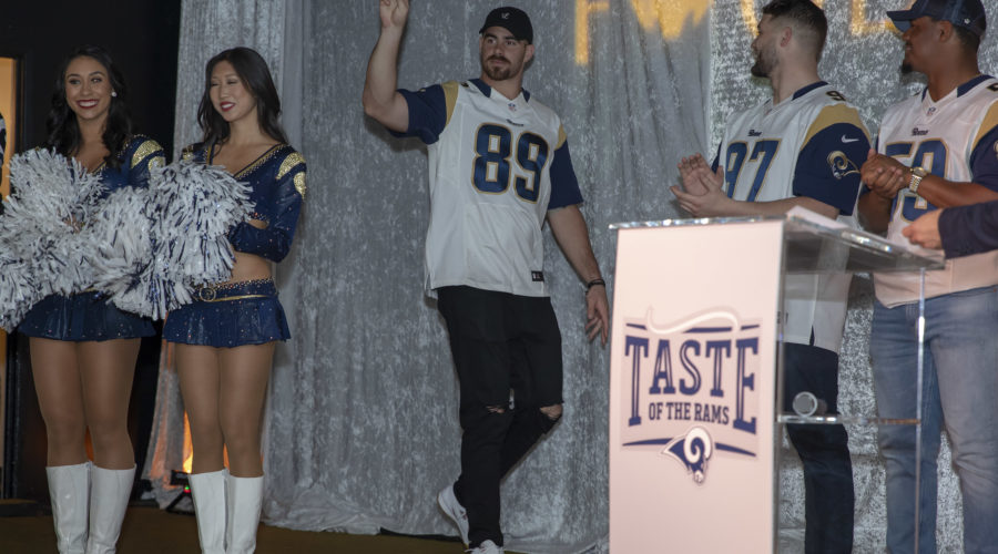 Cheerleaders in blue and silver outfits stand beside a football player in a jersey walking on stage, flashing a peace sign. Two other players and a man in a cap from the Los Angeles Regional Food Bank are at a podium labeled "Taste of the Rams.