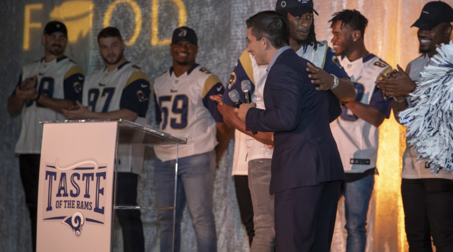 A group of people on stage participates in a "Taste of the Rams" event, with some wearing football jerseys. One person in a suit holds a microphone at the podium, highlighting the Los Angeles Regional Food Bank initiative. The orange-lit backdrop enhances the scene's excitement.