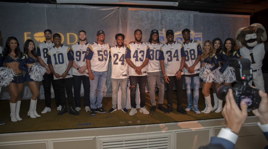 A group of people in matching jerseys and jeans, along with a lively mascot, pose for a photo on stage representing the Los Angeles Regional Food Bank. Cheerleaders in blue outfits stand on either side, while someone captures the moment in the foreground.