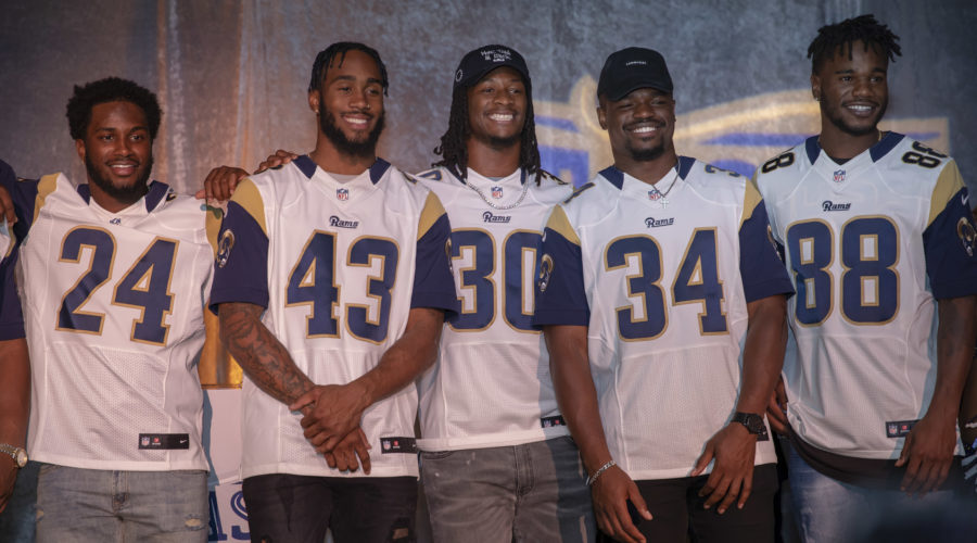 Five individuals are standing in a row, smiling, and proudly wearing white and navy sports jerseys with numbers 24, 43, 30, 34, and 88. They pose in front of a gray backdrop adorned with logos from the Los Angeles Regional Food Bank.