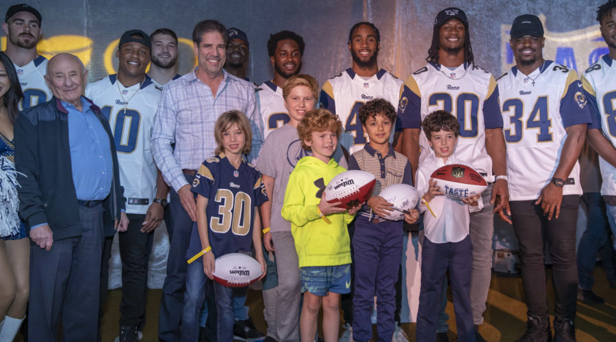 A group of people, including several wearing football jerseys, pose for a photo at what appears to be a sports event supporting the Los Angeles Regional Food Bank. Some hold footballs, and there are both adults and children present, along with a few cheerleaders visible on the side.