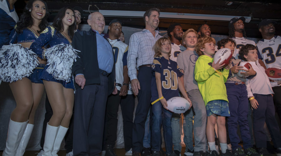 A group of cheerleaders, older men, sports players in uniforms, and children stand together with joy. Some kids hold sports balls as they smile proudly on a stage or platform, united in support of the Los Angeles Regional Food Bank's community initiative.