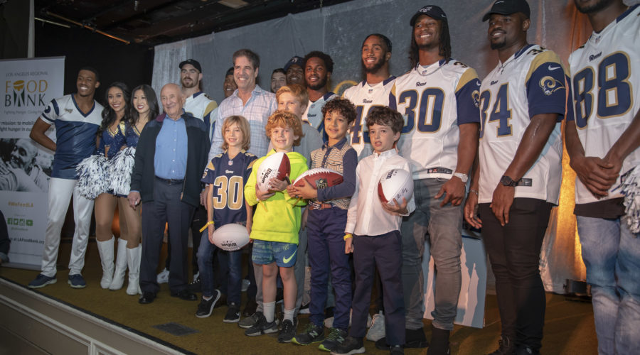 A cheerful group, including cheerleaders, children, and adults in football jerseys, gathers on a stage. Some hold footballs as they stand in front of a Los Angeles Regional Food Bank banner. The mood is lively and friendly.