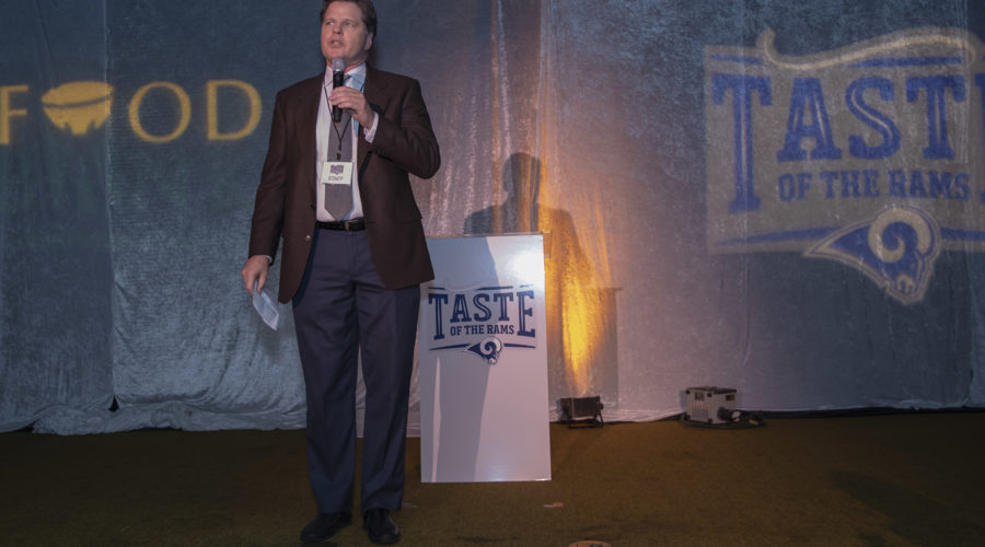 A man in a suit speaks into a microphone on stage at the "Taste of the Rams" event, supporting the Los Angeles Regional Food Bank. The stage backdrop features the event logo and the word "FOOD," while a podium displays the "Taste of the Rams" emblem.