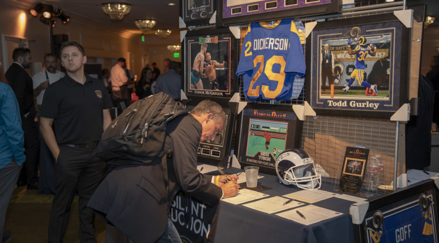 A person is signing a sheet on a table at a memorabilia display supporting the Los Angeles Regional Food Bank. The exhibit features framed jerseys and photos of athletes, including Eric Dickerson and Todd Gurley. Other people are in the background, in a dimly lit room with spotlights.