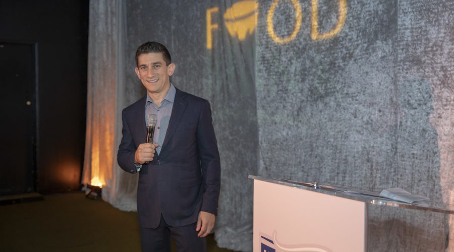 A man in a dark suit stands smiling and holding a microphone on stage. The backdrop features the word "FOOD" partially lit, and a podium with the word "TASTE" before him. This vibrant scene is set at an event for the Los Angeles Regional Food Bank, celebrating their impactful work.