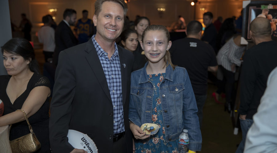At a bustling event for the Los Angeles Regional Food Bank, a man in a suit holds a football beside a smiling young girl in a denim jacket and floral dress. Behind them, people mingle energetically, united by the spirit of giving.