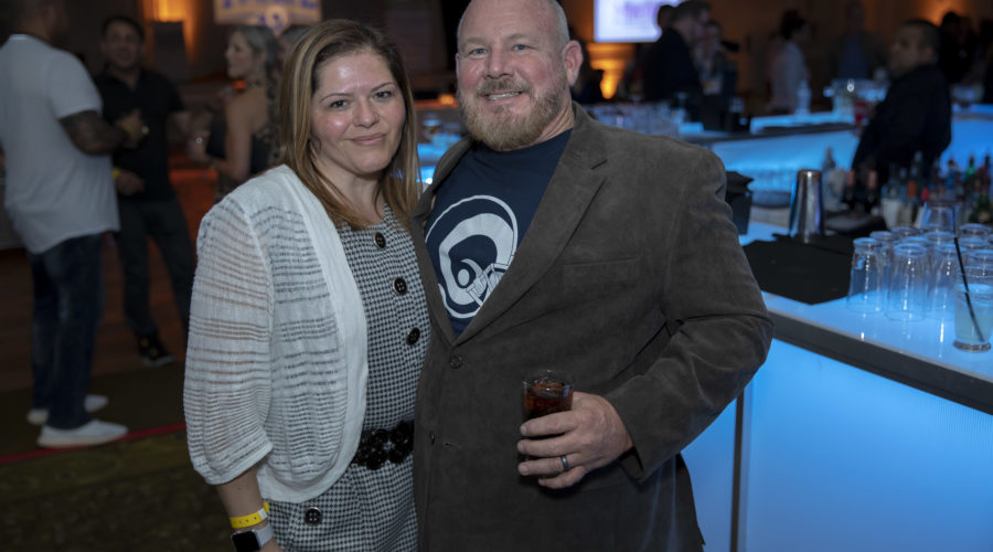 A smiling man and woman pose together at a Los Angeles Regional Food Bank event. The man, holding a drink, sports a blazer over his t-shirt, while the woman dazzles in a patterned outfit. In the background, other guests mingle near the bar.