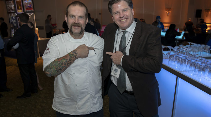 Two men standing and smiling at an event for the Los Angeles Regional Food Bank, one wearing a chef's uniform with tattooed arms, and the other in a suit and tie. Both are pointing at each other in front of a bar area with glasses and dim lighting.