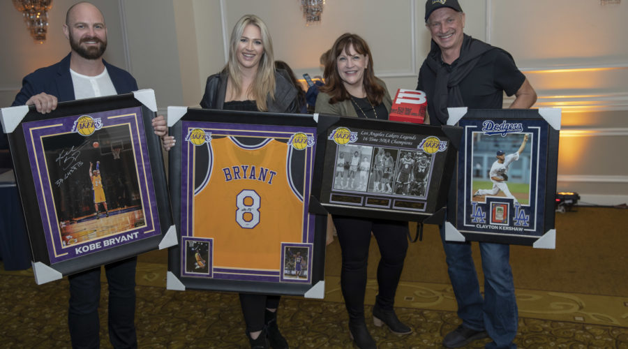 Four people smiling and holding sports memorabilia, including framed items related to Kobe Bryant and Clayton Kershaw. The display, featuring a Lakers jersey and more, takes place in a well-lit room supporting the Los Angeles Regional Food Bank's efforts to combat hunger.