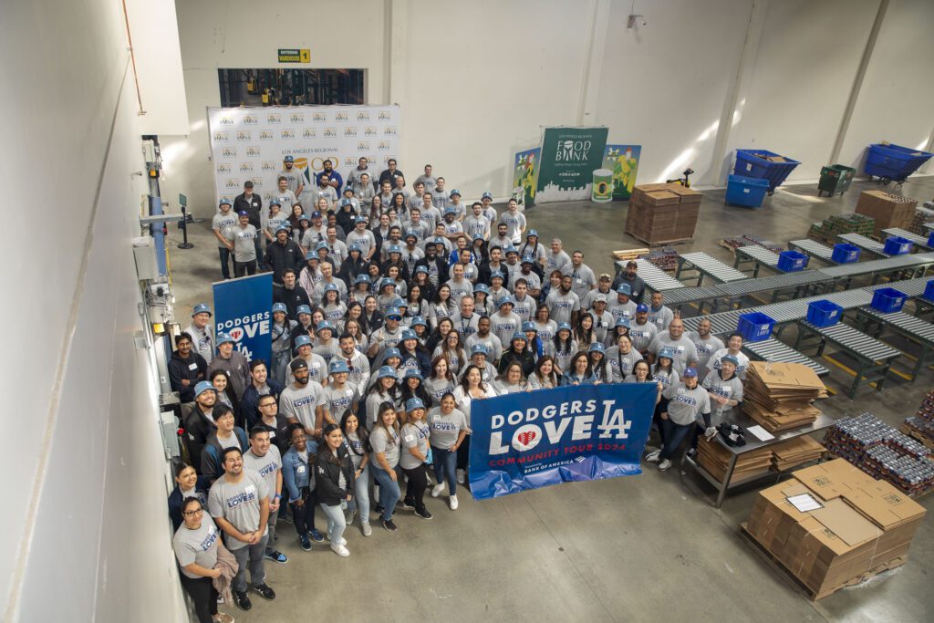 A large group of people wearing gray shirts gathers in a warehouse, smiling and holding a 