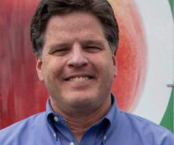 A man with dark hair wearing a blue collared shirt smiles at the camera, standing in front of the Los Angeles Regional Food Bank. A blurred image of a large fruit can be seen in the background, highlighting his commitment to nourishing communities.