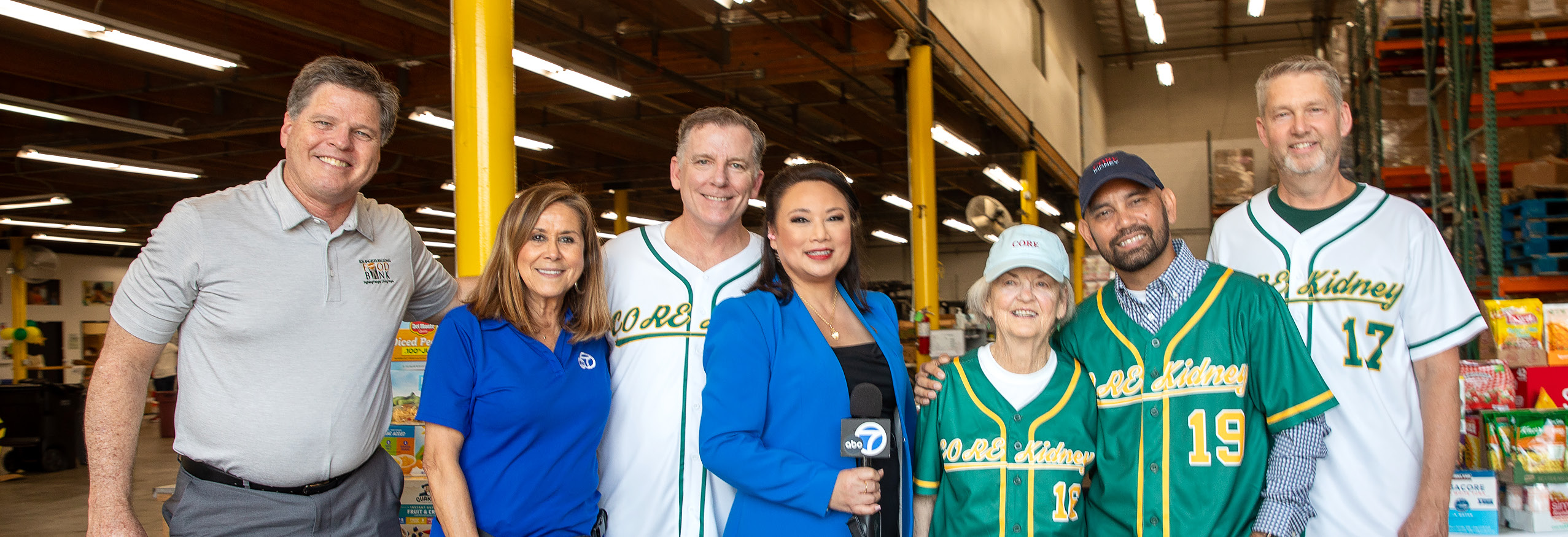 A group of eight people, smiling in a warehouse at the Los Angeles Regional Food Bank. Some wear matching baseball jerseys as one holds a microphone, ready to inspire. Shelves filled with boxes create a bustling backdrop for this lively team.