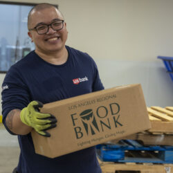 A smiling person with glasses and green gloves, representing the spirit of US Bank, holds a cardboard box labeled "Los Angeles Regional Food Bank" in a bustling warehouse. Wooden pallets and recycling bins fill the background, showcasing the commitment to community support.