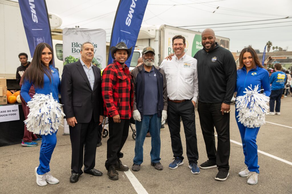 A group of six people, four men in casual and business attire and two women in blue cheerleader uniforms, pose together outdoors at a food bank event. Some hold white pom-poms. Banners and a truck are visible in the background.