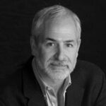 Black and white photo of an older man with short white hair and a beard. He is wearing a dark blazer over a light shirt, looking directly at the camera with a neutral expression against a dark background, evoking the dignity and resilience found in those who support food banks.