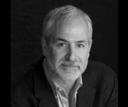 A grayscale photo of an older man with short hair and a beard, wearing a dark suit jacket over a light shirt. He gazes directly at the camera with a neutral expression, as if contemplating the vital work of a food bank, all against a plain dark background.