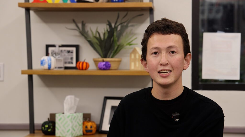 A person with short brown hair, wearing a black shirt and a small microphone, sits indoors in front of shelves decorated with framed photos, potted plants, a tissue box, small Halloween pumpkins, and food pantry essentials.