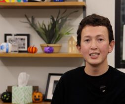 A person with short brown hair, wearing a black shirt and a small microphone, sits indoors in front of shelves decorated with framed photos, potted plants, a tissue box, small Halloween pumpkins, and food pantry essentials.
