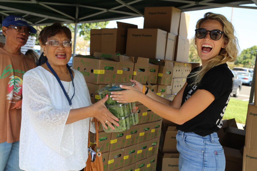 Two women smile as they hold a plastic container of cucumbers together at an outdoor food bank distribution event, with boxes stacked in the background and a man standing nearby.
