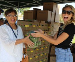 Two women smile as they hold a plastic container of cucumbers together at an outdoor food bank distribution event, with boxes stacked in the background and a man standing nearby.