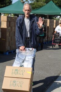 An older man waves while pushing a cart loaded with food bank boxes outdoors. Rows of cardboard boxes and green tents are visible in the background at a food bank distribution event.