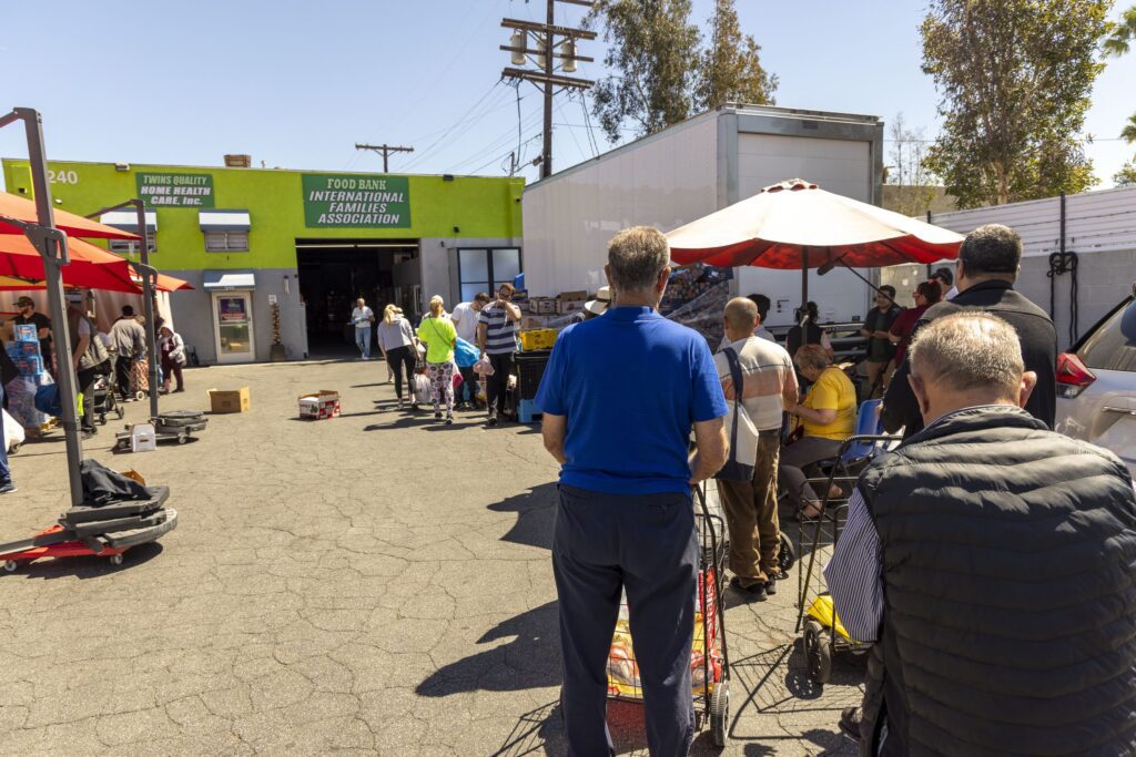 People stand in a line outside a food pantry under sunny skies. Red umbrellas provide shade, and volunteers assist at tables. The atmosphere appears orderly as people wait for assistance.