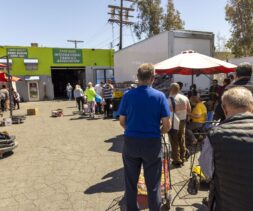 People stand in a line outside a food pantry under sunny skies. Red umbrellas provide shade, and volunteers assist at tables. The atmosphere appears orderly as people wait for assistance.