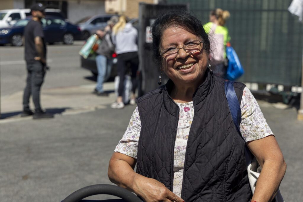 An older woman wearing glasses, a black vest, and a floral shirt smiles outdoors on a sunny day near a local food pantry. Several people and cars are visible in the blurred background.