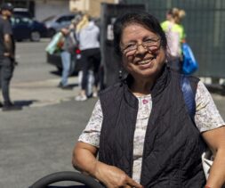 An older woman wearing glasses, a black vest, and a floral shirt smiles outdoors on a sunny day near a local food pantry. Several people and cars are visible in the blurred background.