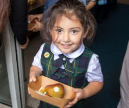 A young girl in a plaid school uniform holds a tray with a pear and a small packet, smiling brightly while standing indoors near a doorway—capturing the positive spirit highlighted in our recent Impact Report.