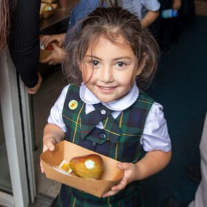 A young girl in a plaid school uniform holds a tray with a pear and a small packet, smiling brightly while standing indoors near a doorway—capturing the positive spirit highlighted in our recent Impact Report.