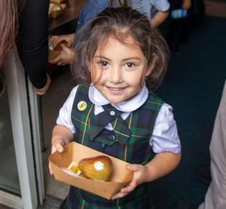 A young girl in a plaid school uniform holds a tray with a pear and a small packet, smiling brightly while standing indoors near a doorway—capturing the positive spirit highlighted in our recent Impact Report.