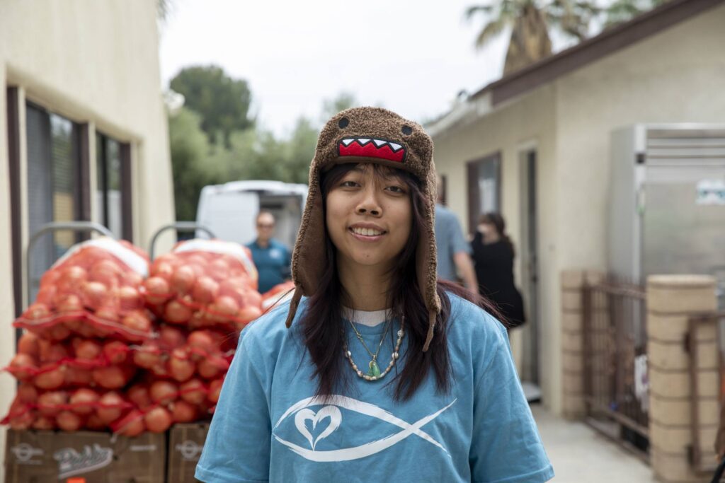 Young person wearing a brown animal hat and blue shirt with a heart and fish symbol stands outdoors near bags of tomatoes, part of a food assistance event. Other people and buildings are visible in the background.