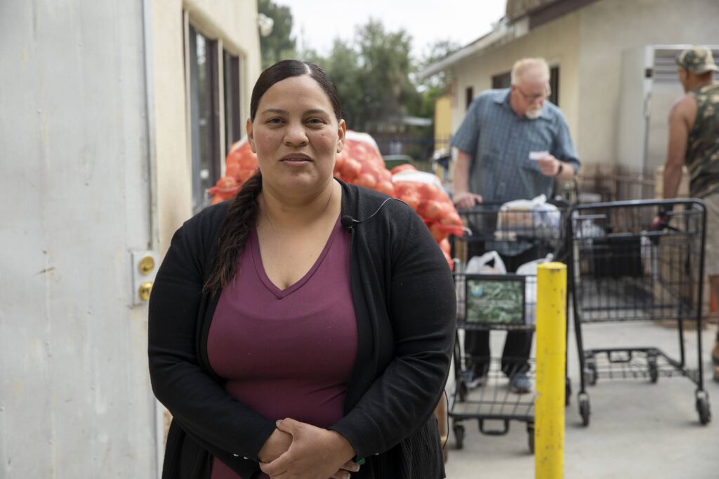 A woman volunteer stands outside, smiling at the camera, with bags of produce and shopping carts behind her. People in the background appear to be collecting groceries in an outdoor setting.