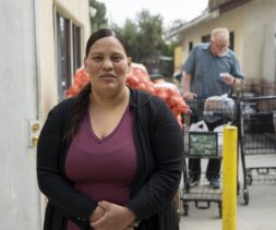 A woman volunteer stands outside, smiling at the camera, with bags of produce and shopping carts behind her. People in the background appear to be collecting groceries in an outdoor setting.