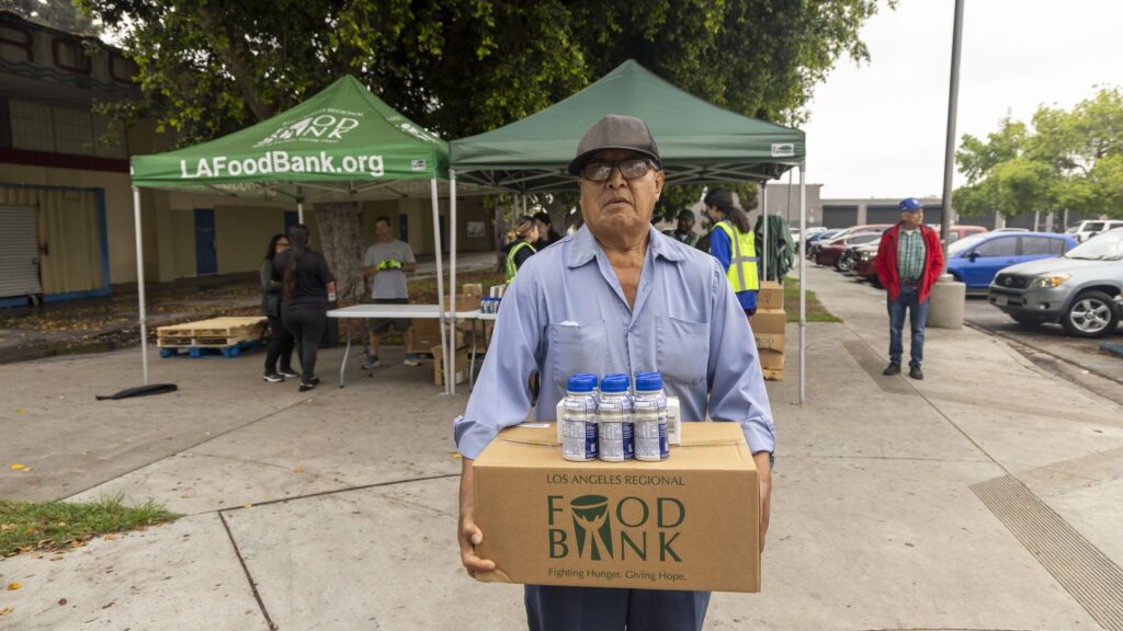An older man wearing a cap and glasses carries a box labeled 