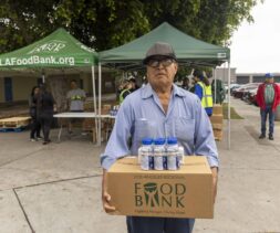An older man wearing a cap and glasses carries a box labeled 