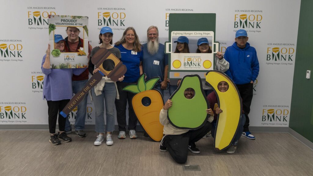 A group of people pose cheerfully in front of a Los Angeles Regional Food Bank wall, holding fruit-shaped cutouts, volunteer signs, and food from the bar, celebrating community service and fighting hunger.