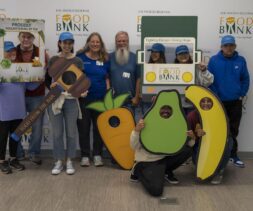 A group of people pose cheerfully in front of a Los Angeles Regional Food Bank wall, holding fruit-shaped cutouts, volunteer signs, and food from the bar, celebrating community service and fighting hunger.