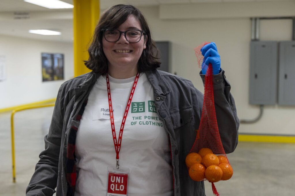 A person wearing glasses and a UNIQLO badge smiles while holding a mesh bag of small oranges, ready to help at the food bank. They are indoors in a white UNIQLO t-shirt, gray jacket, and one blue glove.